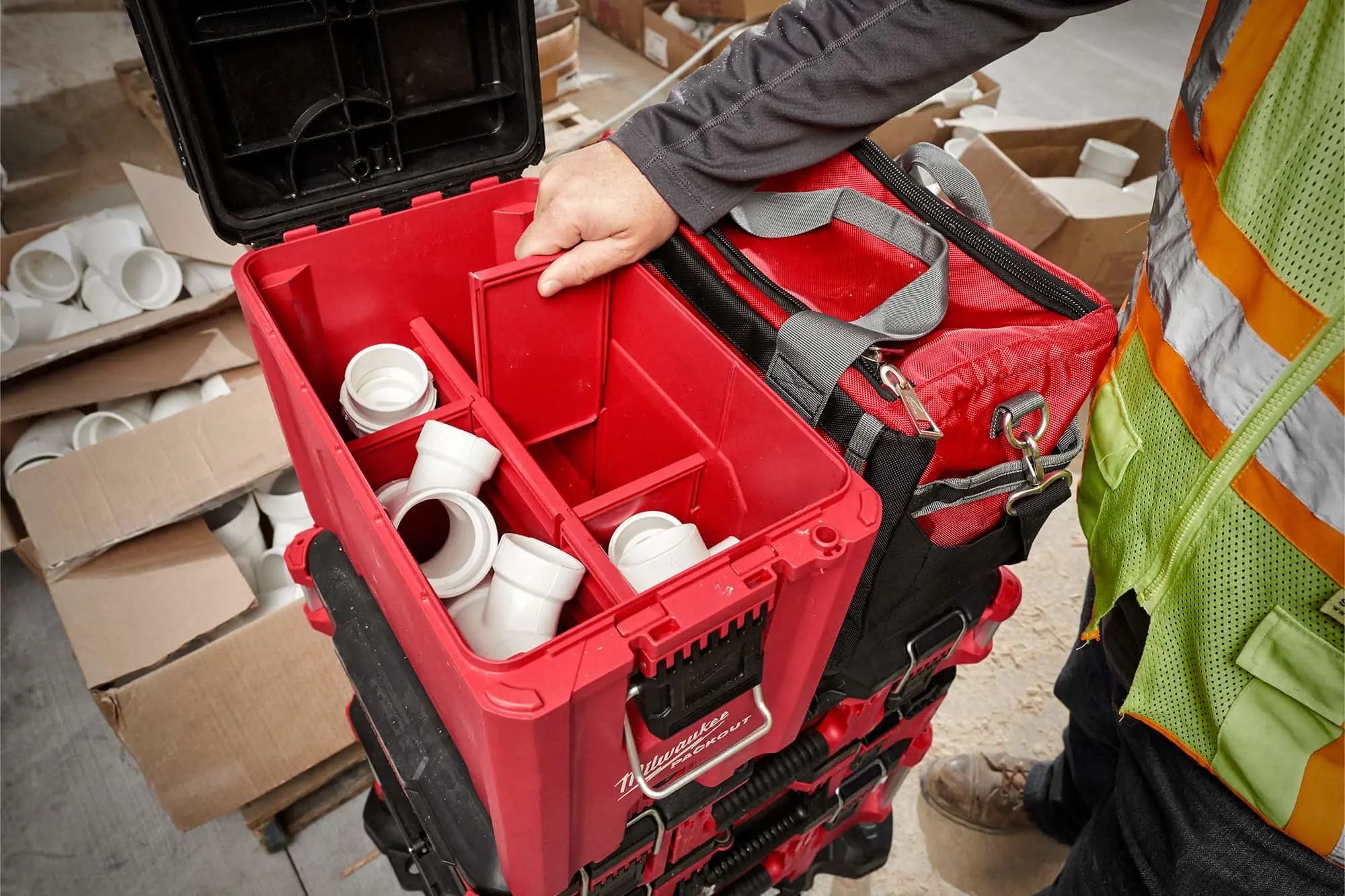 Person opening red Milwaukee toolbox containing white plumbing fittings, with tool bag and cardboard boxes nearby.