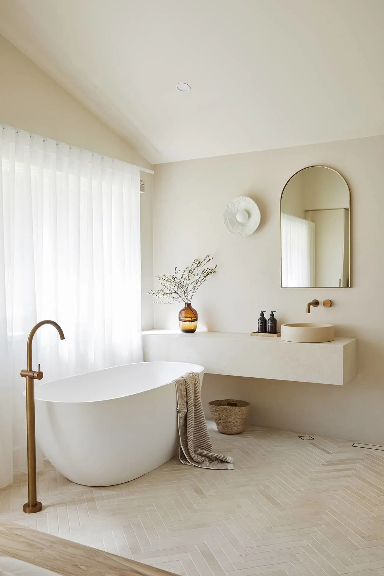 Modern minimalist bathroom with white freestanding tub, brass fixtures, floating sink, and herringbone tile floor.