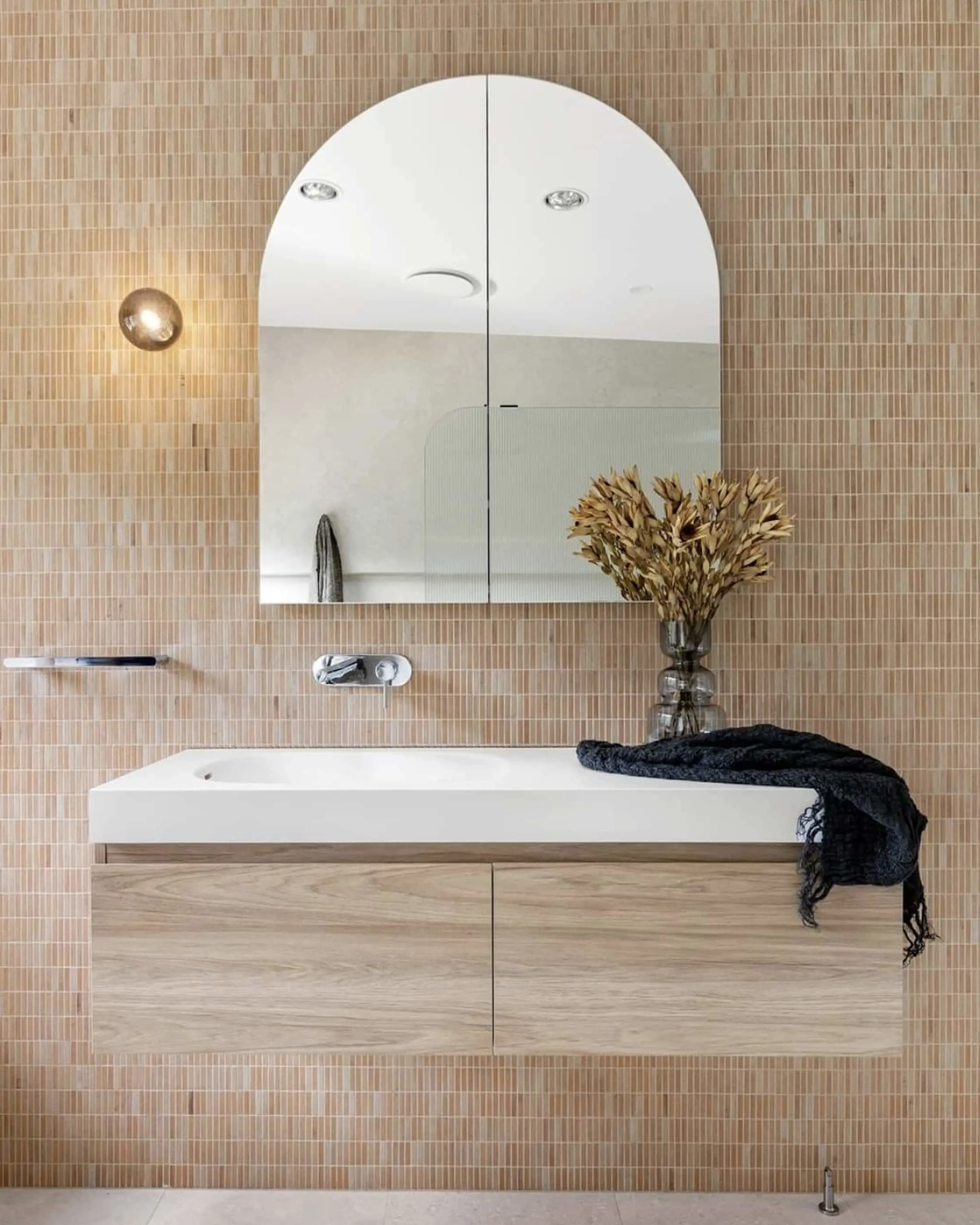 Modern bathroom with peach tile walls, arch mirror, white sink on wood vanity, dried flowers in vase, and black towel.