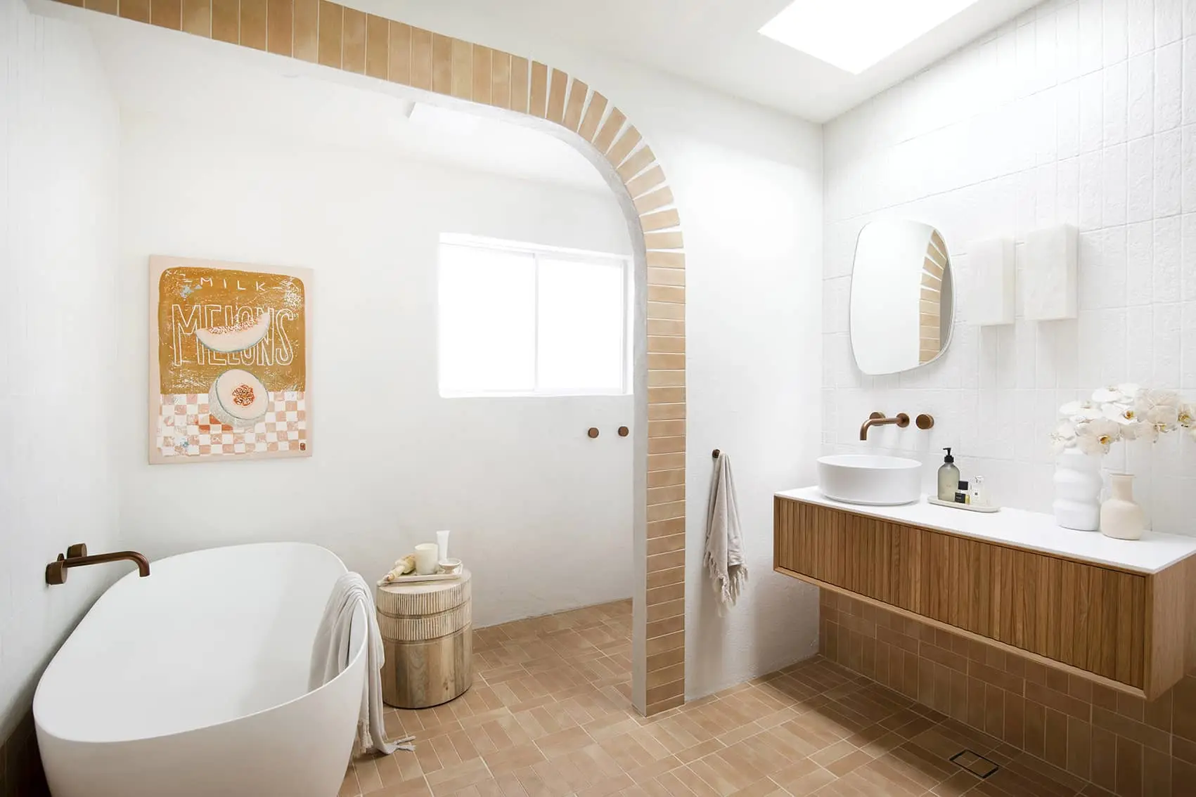 Modern white bathroom with freestanding tub, wooden vanity, terracotta tile floor, and arched doorway with decorative wall art.