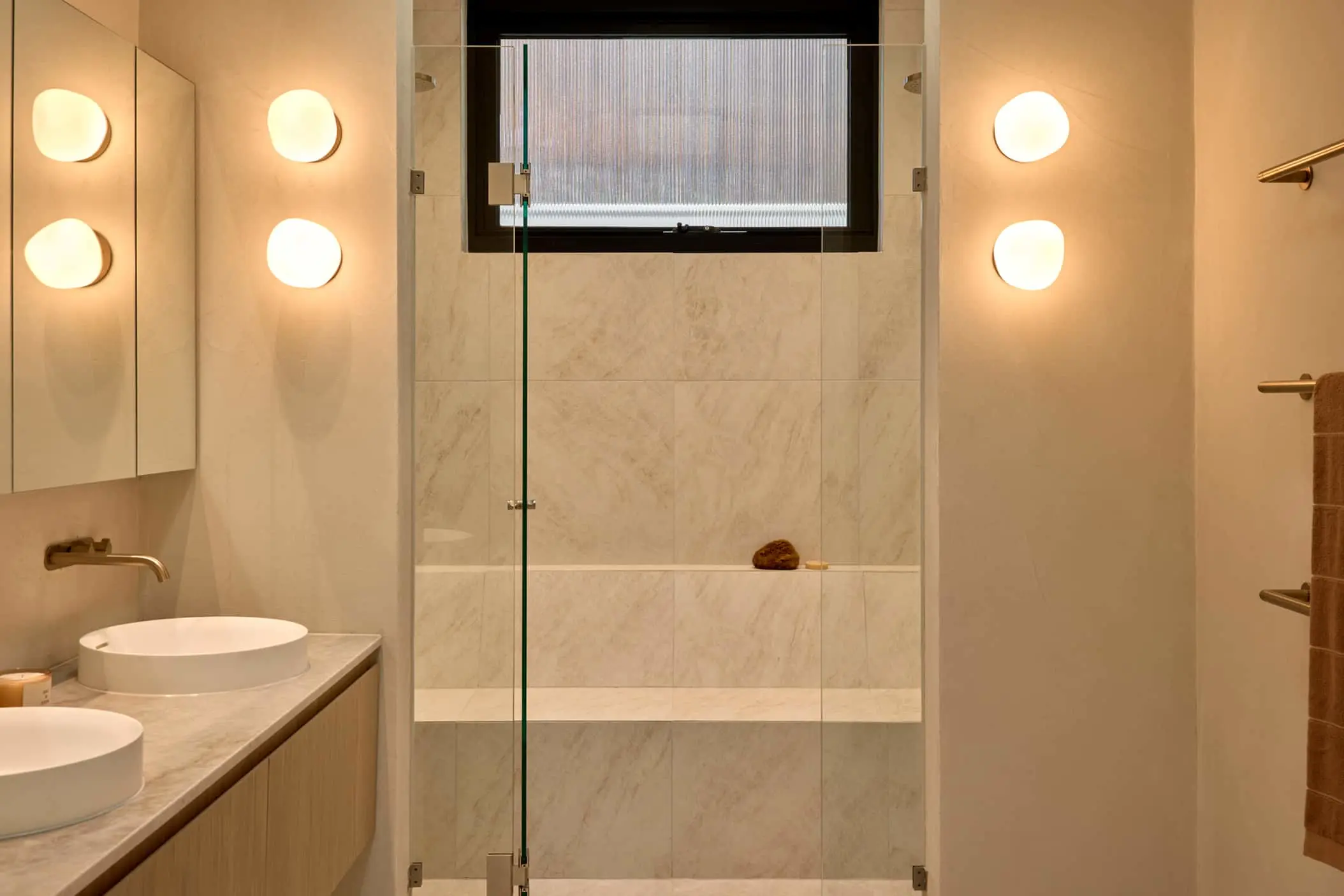 Modern bathroom with beige marble tiles, glass shower enclosure, round wall lights, and white vessel sinks.