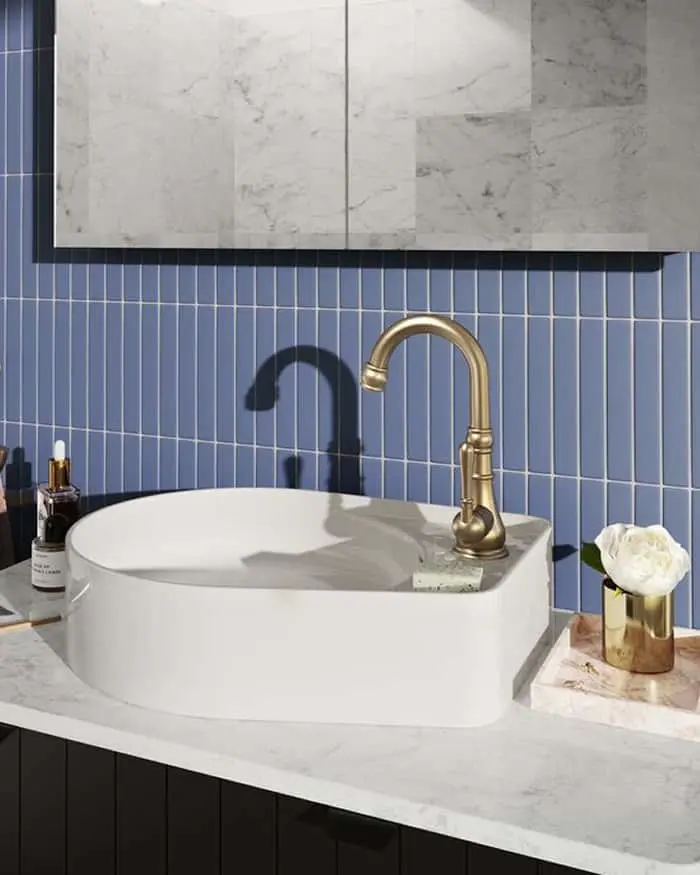 Modern bathroom with white vessel sink, brass faucet, blue tile backsplash, marble countertop, and small white flower in gold vase.