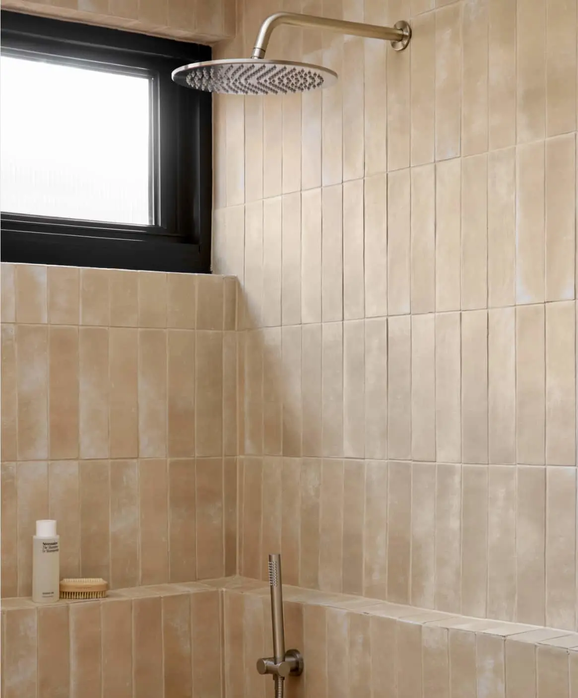 Modern bathroom with beige vertical tiles, round rainfall showerhead, black-framed window, and built-in shelf with toiletries.