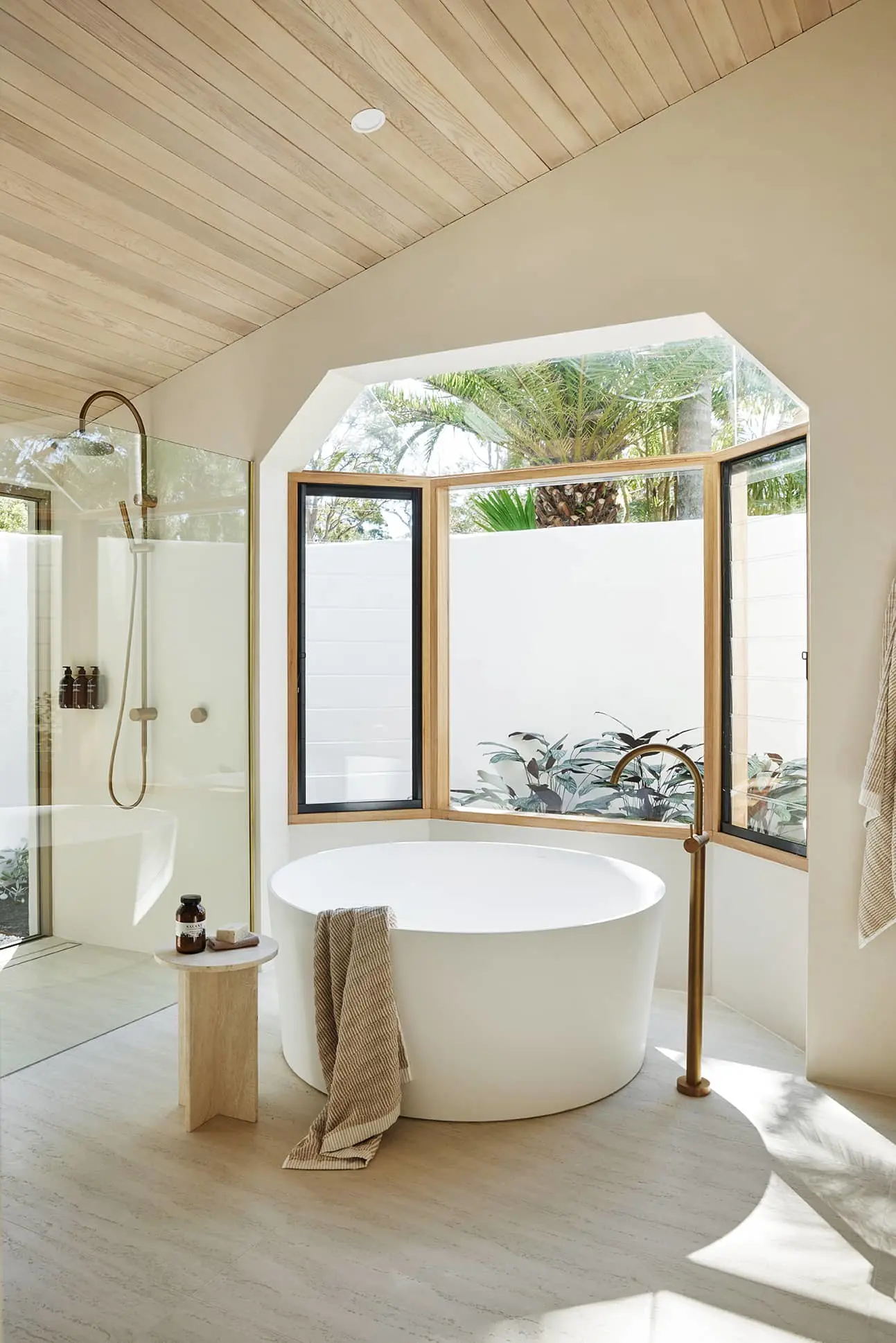 Modern bathroom with round white bathtub, glass shower, and large windows overlooking tropical plants and palm trees.