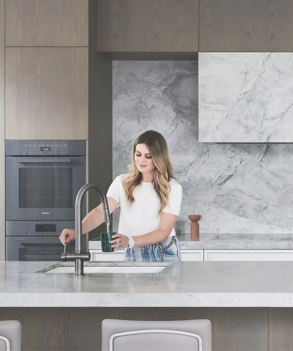 Person filling glass at kitchen sink in modern kitchen with marble backsplash, wooden cabinets and built-in oven.