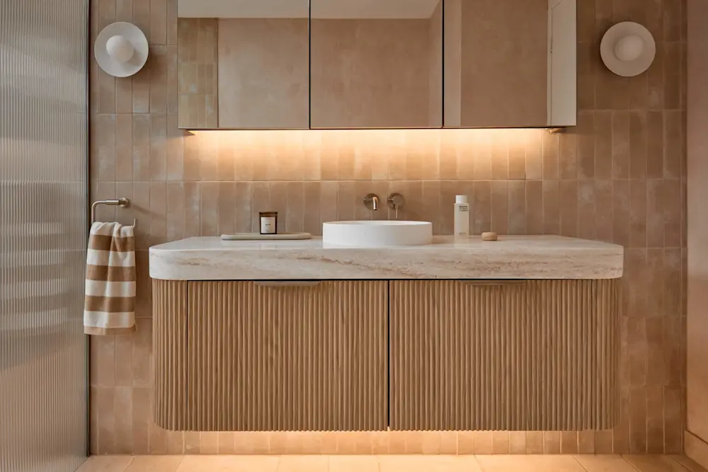 Modern bathroom with beige tiled walls, wooden vanity with stone countertop, white vessel sink, and under-cabinet lighting.