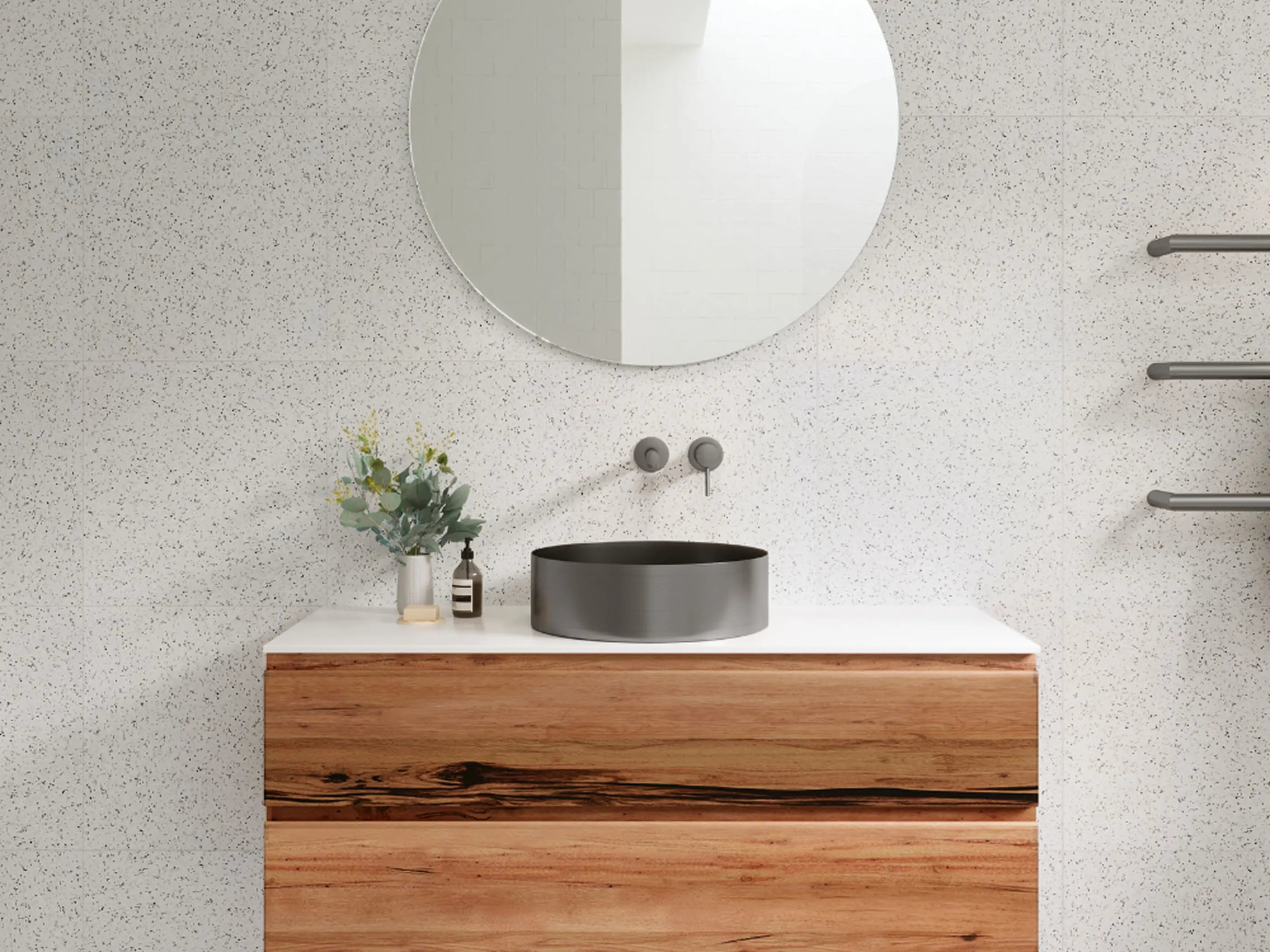 Modern bathroom with wooden vanity, black circular sink, round mirror, and small plant against speckled white walls.