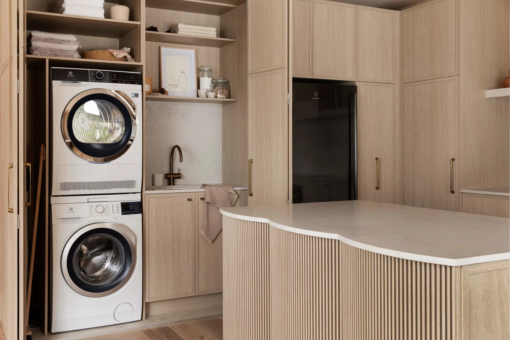 Modern laundry room with stacked washer and dryer, light wood cabinetry, small sink, and central island counter.