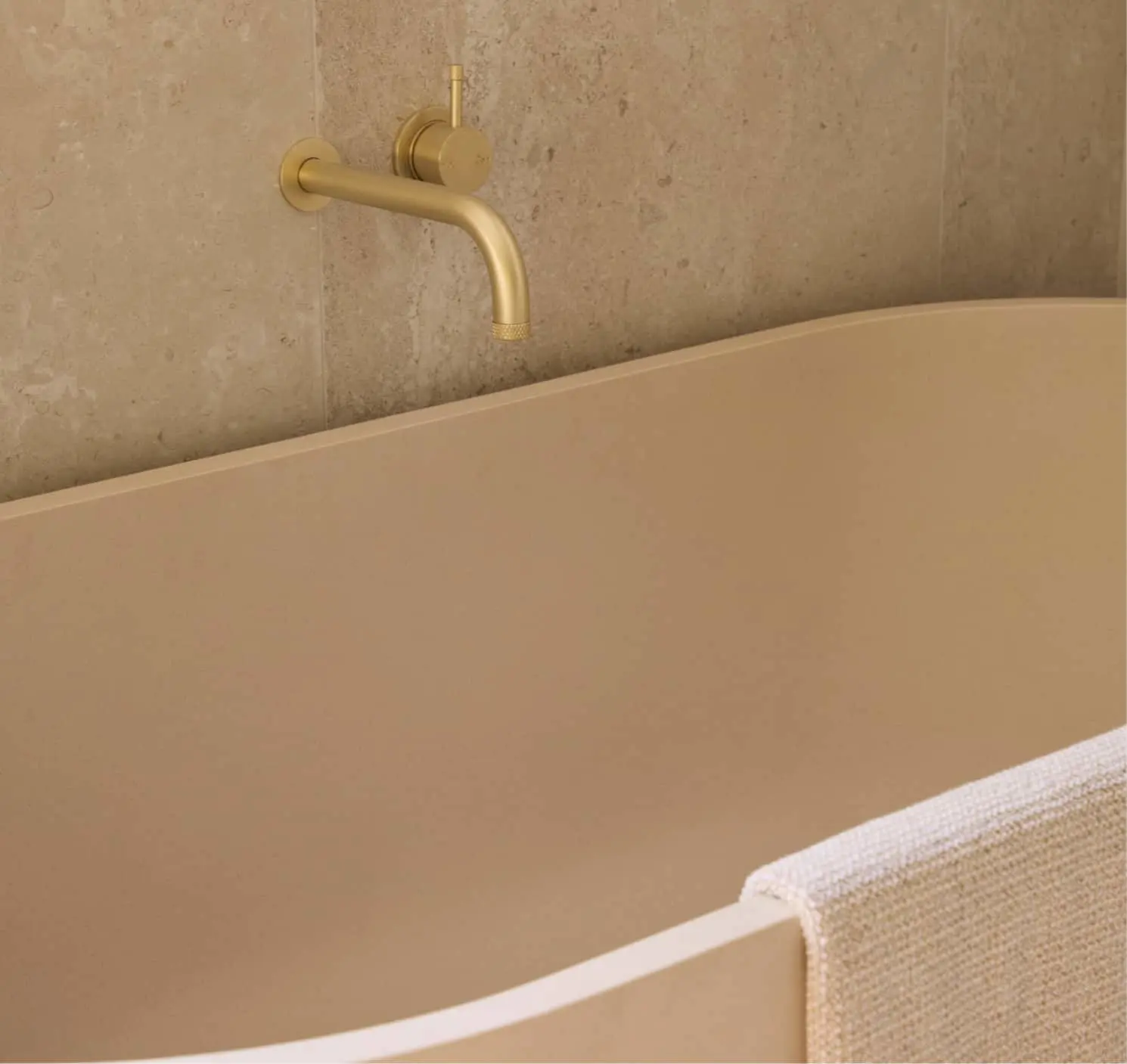 A beige freestanding bathtub with a gold wall-mounted faucet against a travertine stone wall, with a white towel draped over the tub edge.