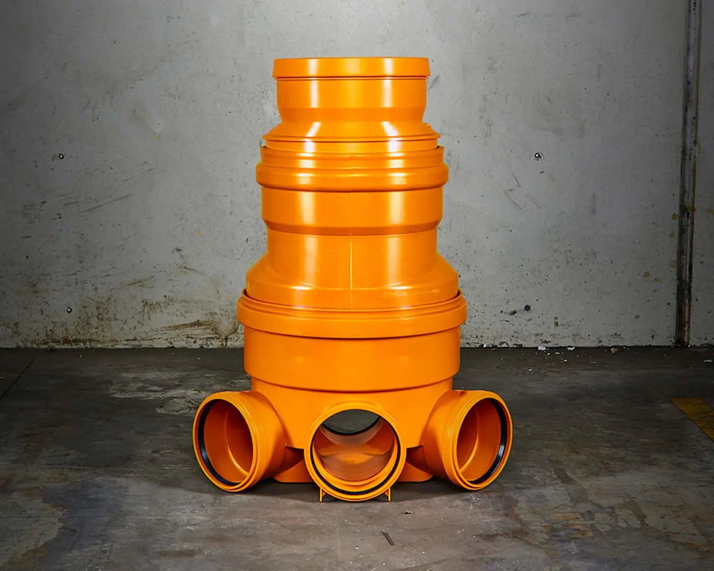Bright orange plastic plumbing fixture with multiple pipe connections against a concrete wall.