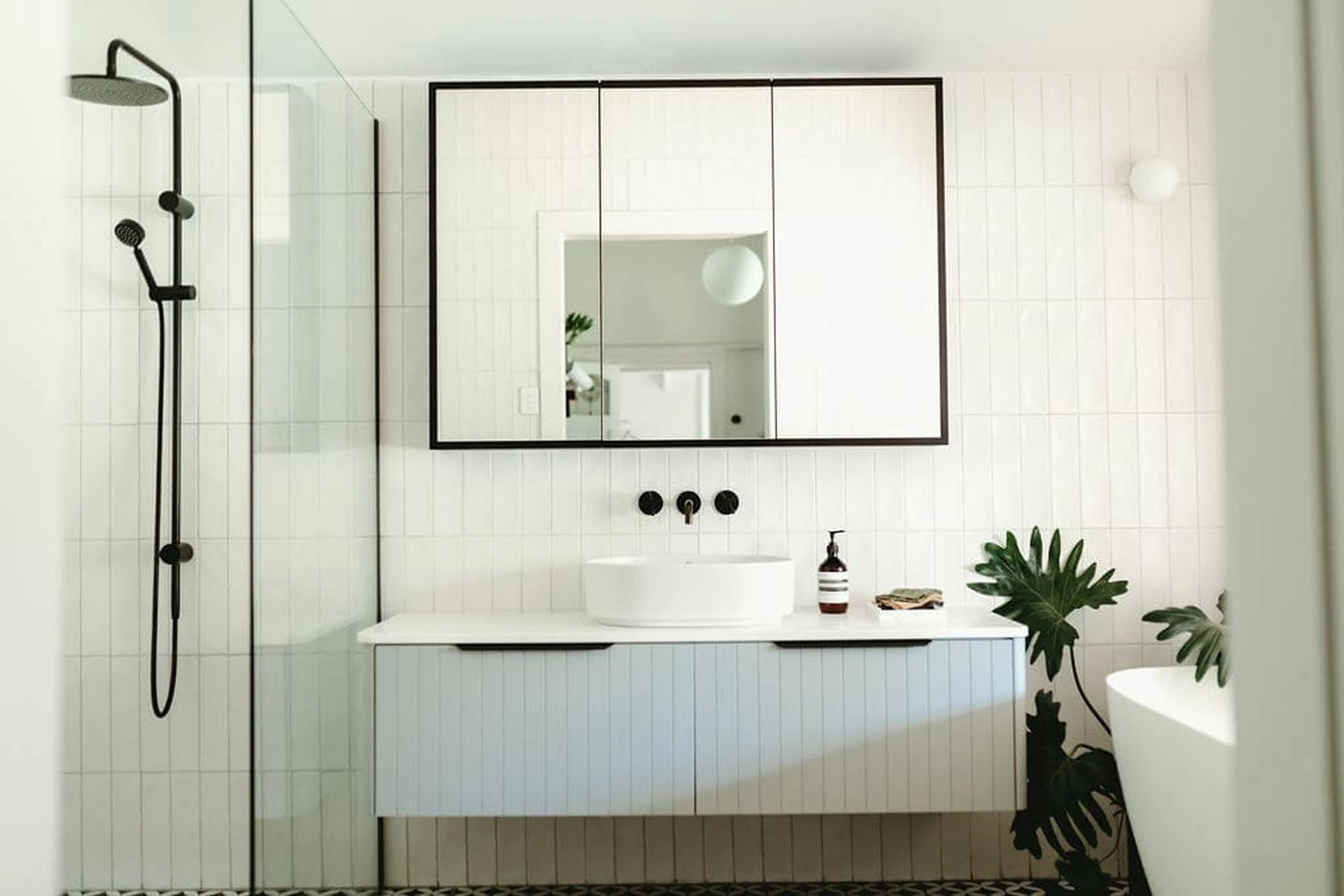 Modern white bathroom with black-framed mirror, vessel sink, glass shower, and green plant in corner.