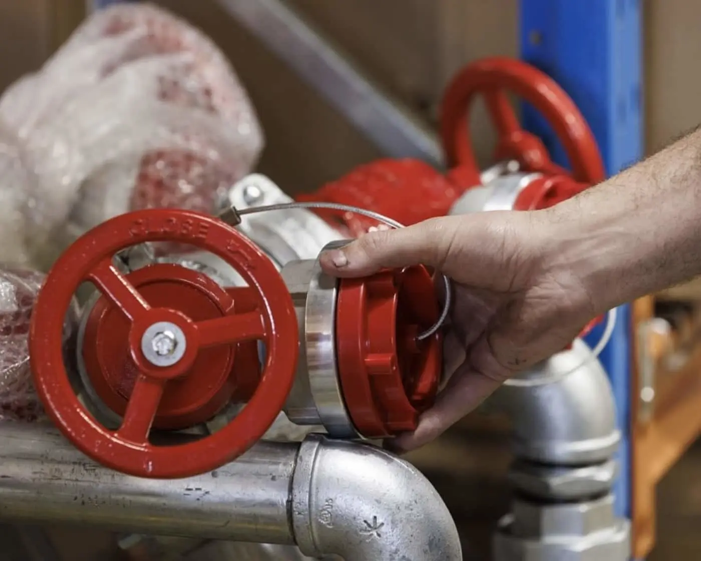 Hand turning a red valve wheel on industrial piping, with multiple valves visible on metal pipes.