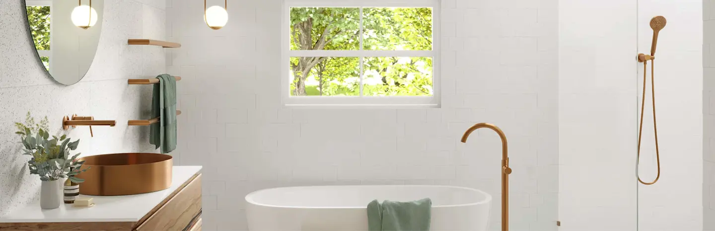 Modern white bathroom with bronze sink, freestanding tub, brass fixtures, and window overlooking green trees.