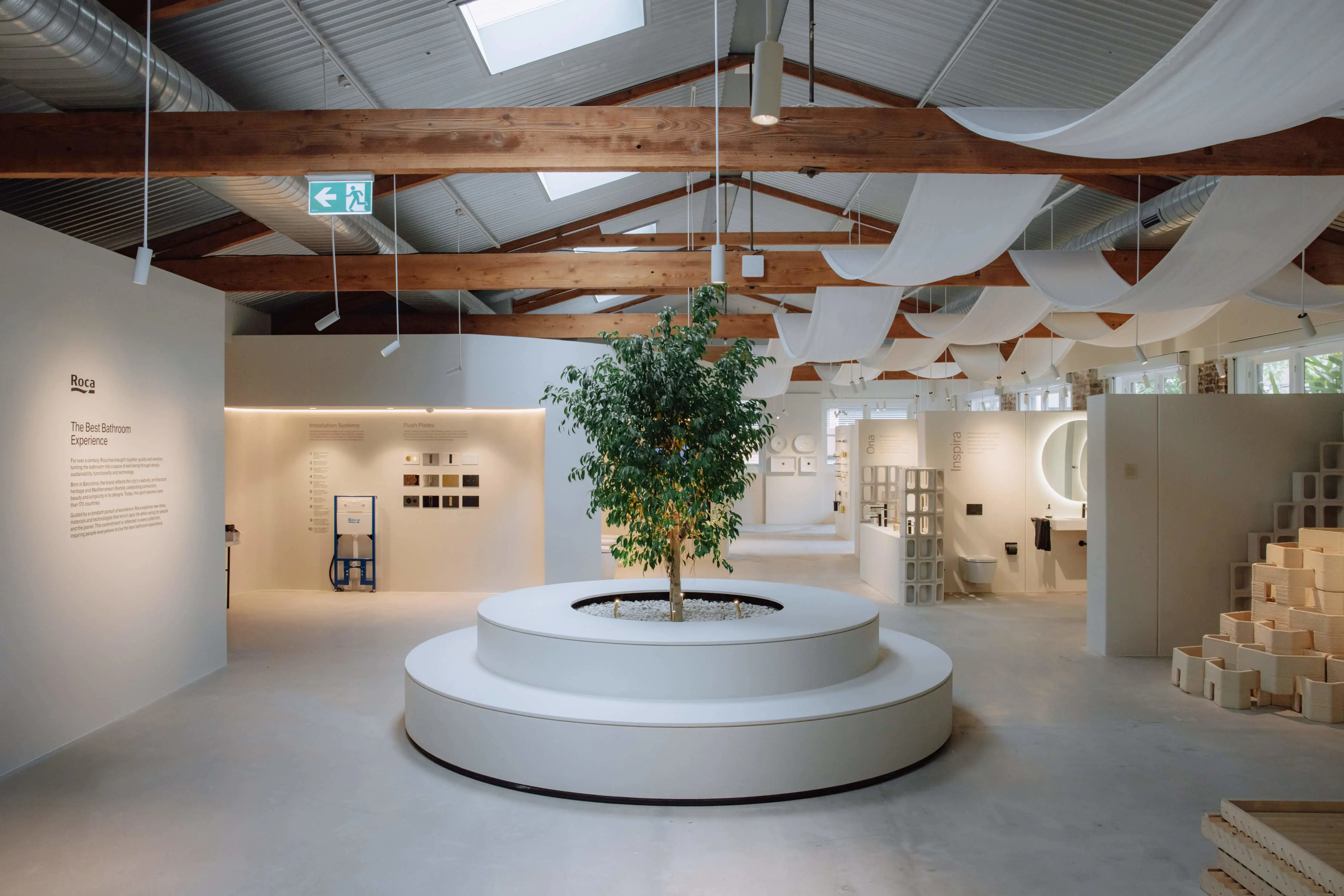 Modern bathroom showroom with central tree display, wooden ceiling beams, white walls, and bathroom fixtures on display.