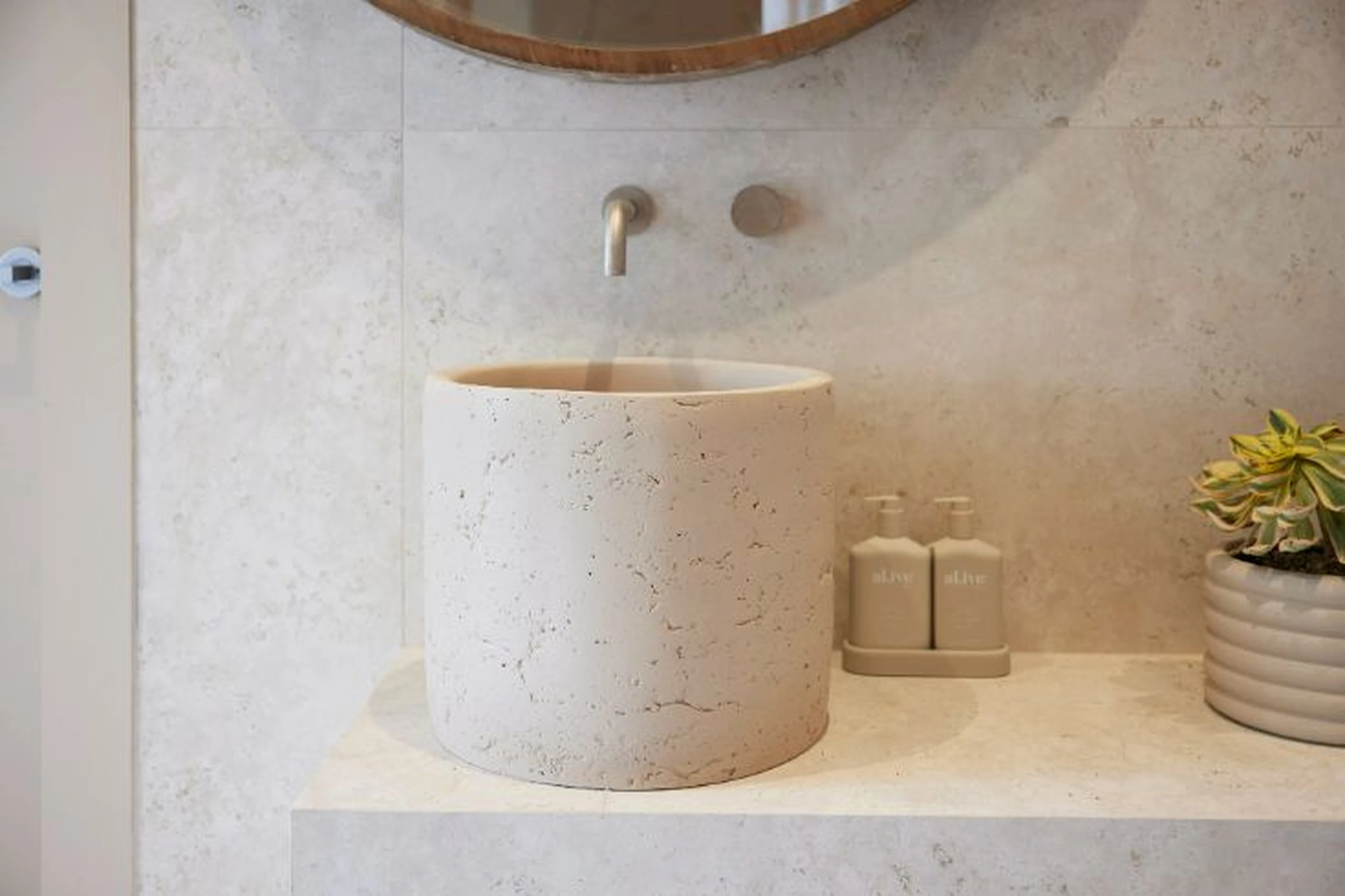 Modern bathroom with cylindrical stone sink, wall-mounted faucet, cream dispensers, and small plant in neutral tones.