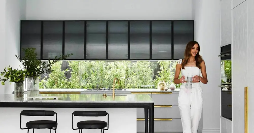 Woman in white outfit standing in modern kitchen with black countertops, gold fixtures, and garden window view.