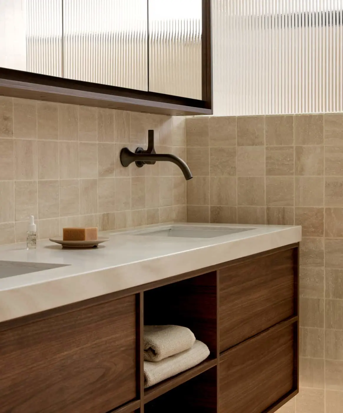 Modern bathroom vanity with white countertop, dark wood cabinets, wall-mounted bronze faucet, and beige tiled walls.