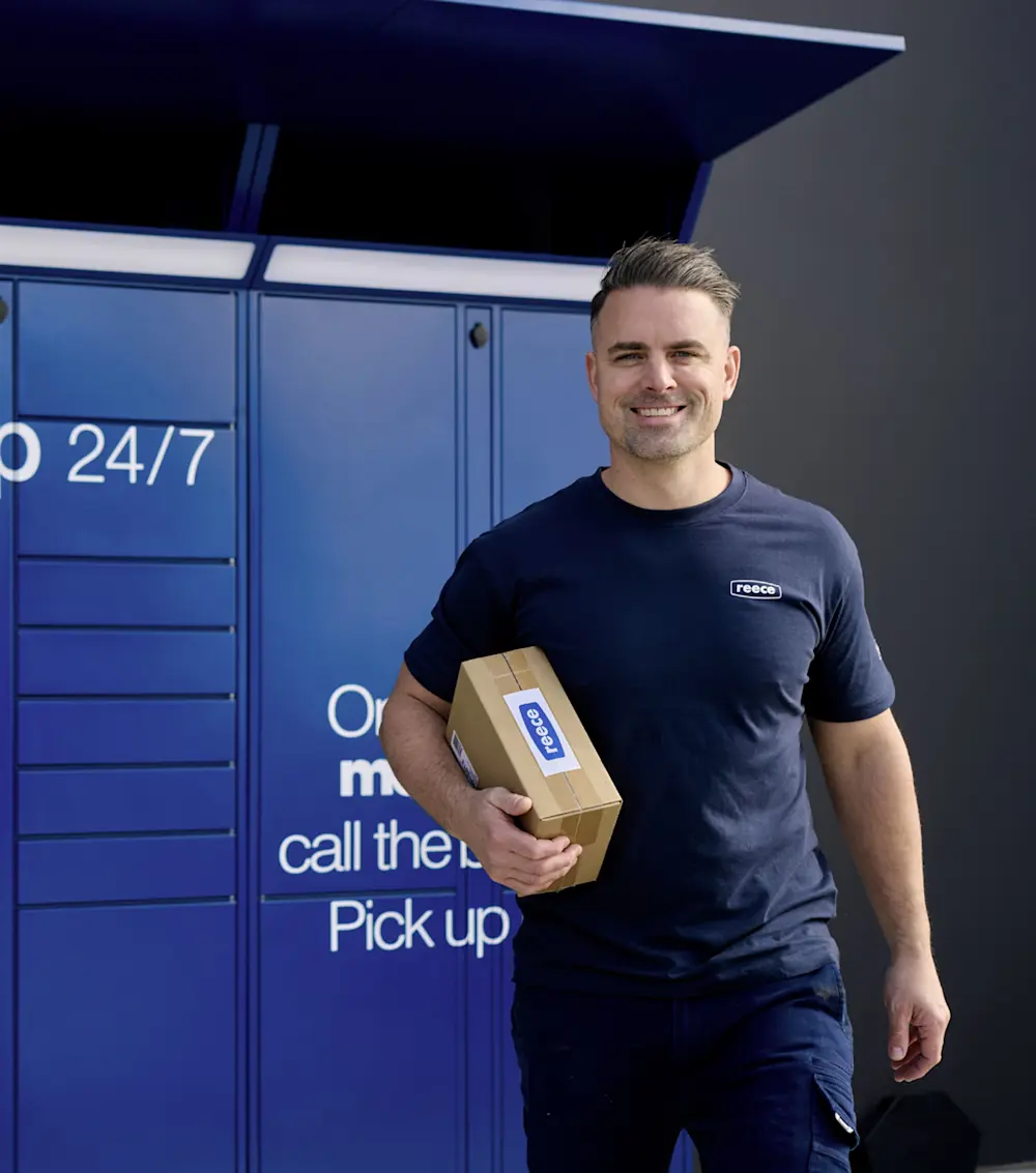 Person in navy Reece uniform holding a package in front of blue parcel lockers with "24/7" visible.