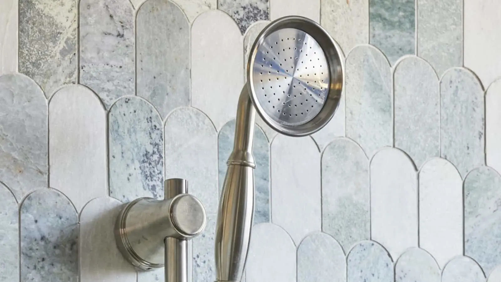 Stainless steel handheld shower head mounted against light blue and white marble tile backsplash with scalloped pattern.
