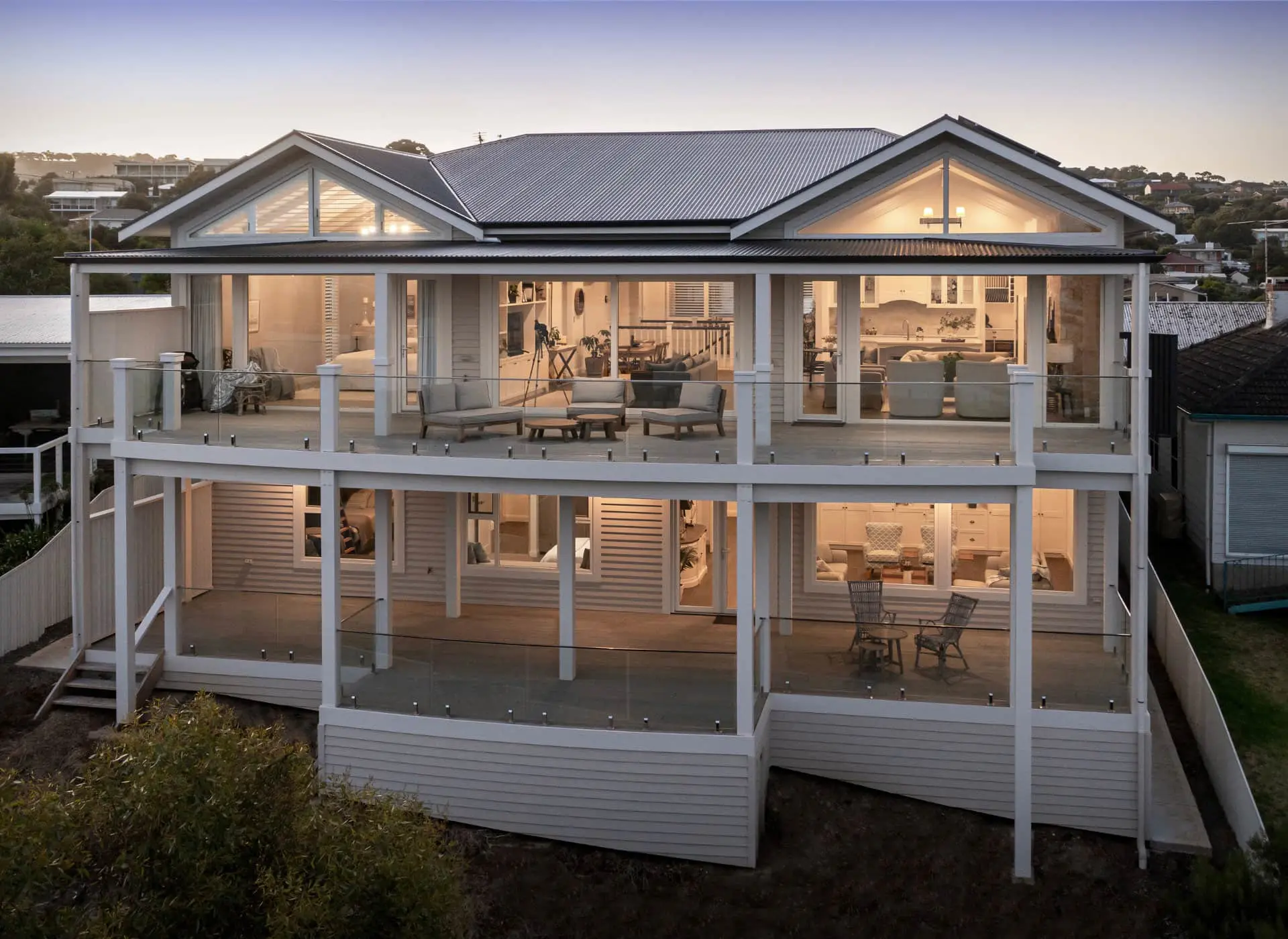 Modern two-story white house with glass balconies and large windows illuminated at dusk, showing interior living spaces.