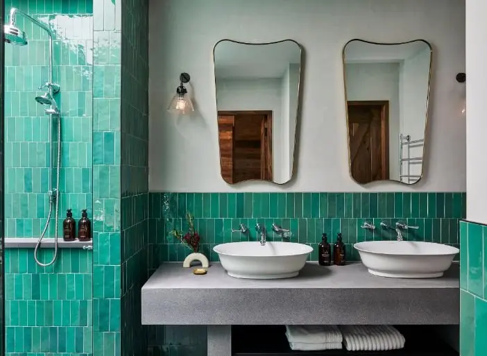 Modern bathroom with turquoise tile, glass shower, double white vessel sinks, and two framed mirrors above gray counter.