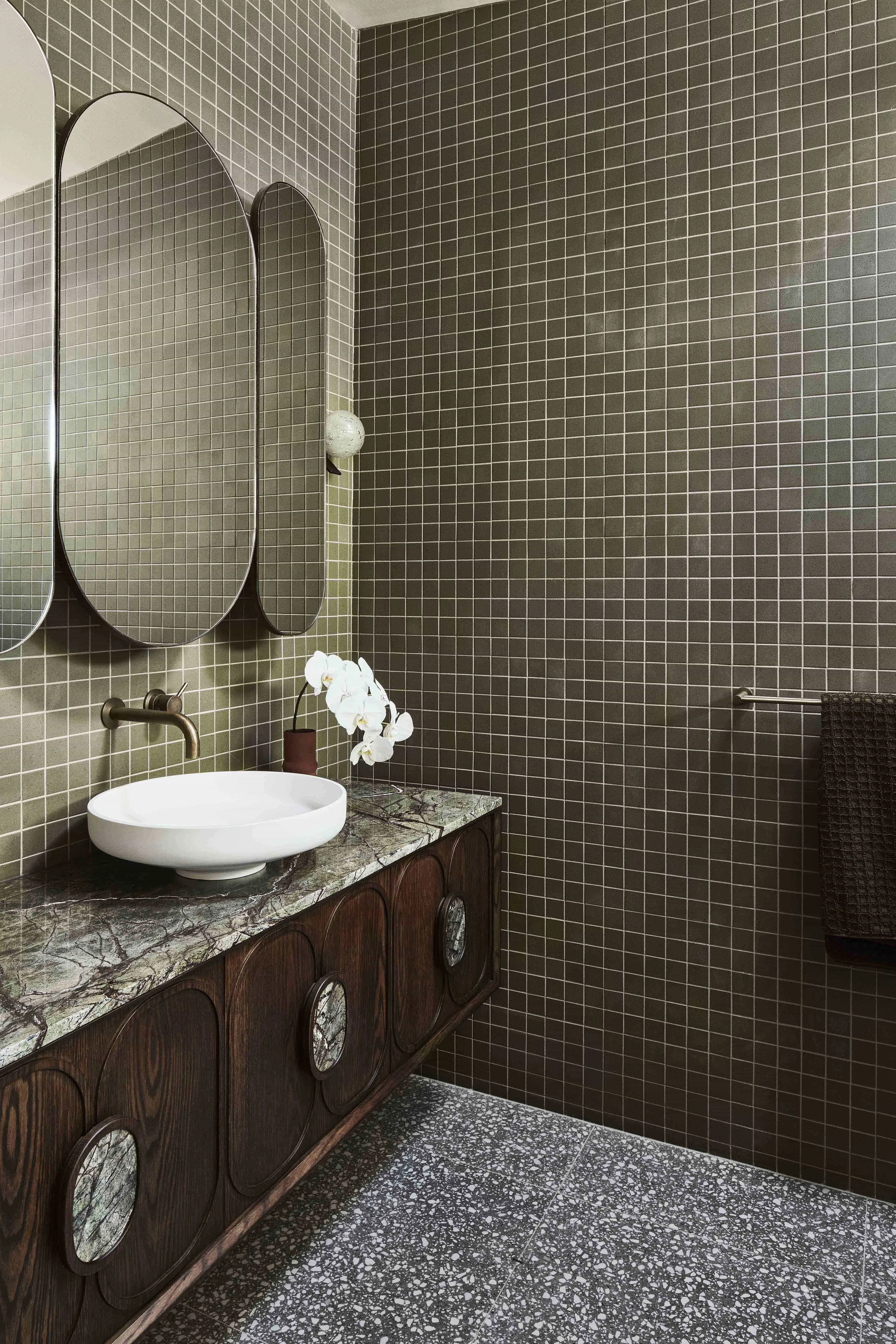 Bathroom with dark green square tiles, terrazzo floor, wood vanity with marble top, white vessel sink, oval mirrors, and white orchid.