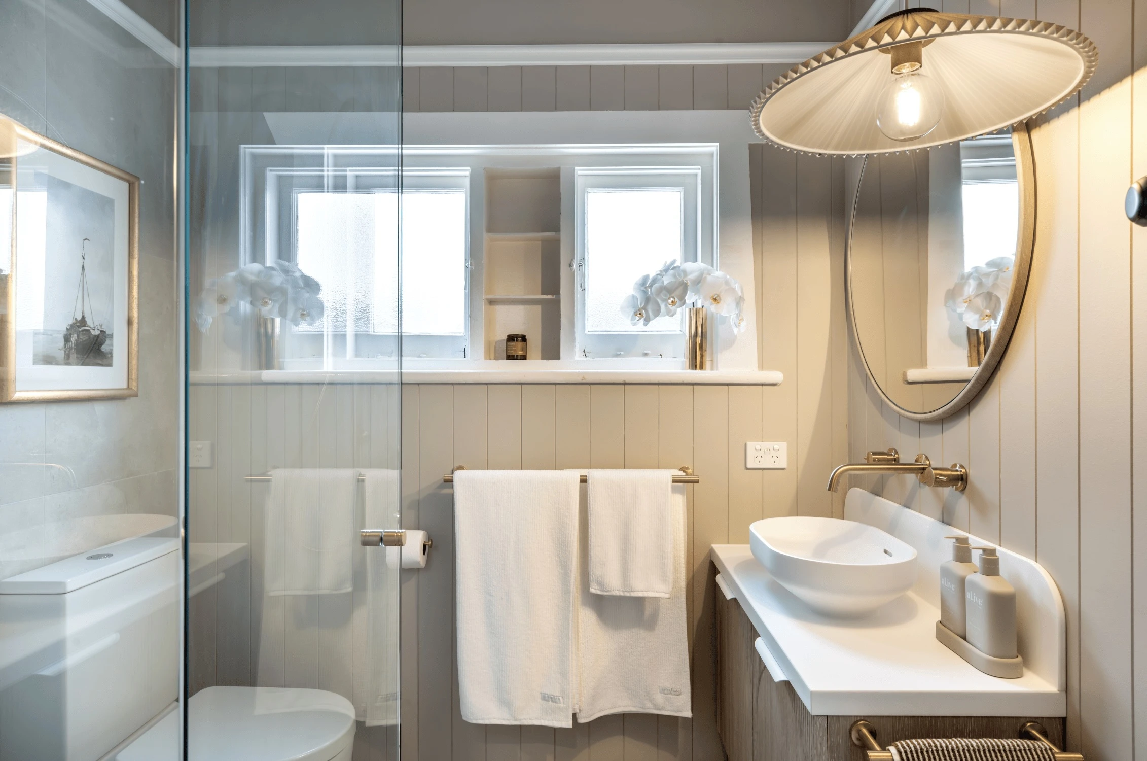 Modern bathroom with white paneled walls, glass shower, round mirror, pendant light, and white orchids on windowsill.