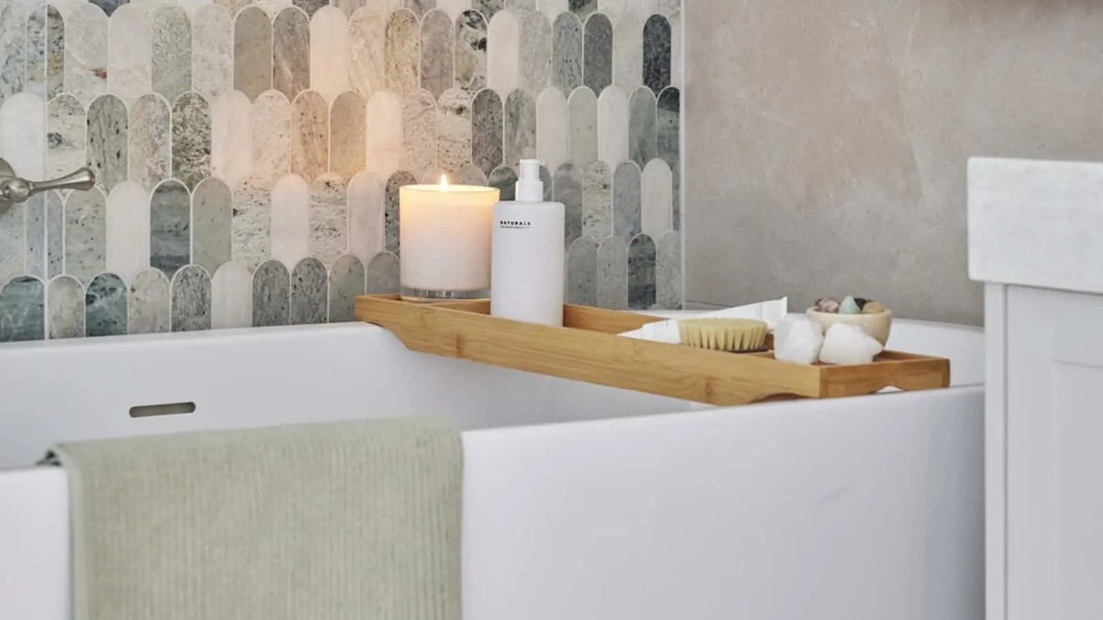 Elegant bathroom with wooden tray on white bathtub holding lit candle, lotion bottle, and bath accessories against marble tile.
