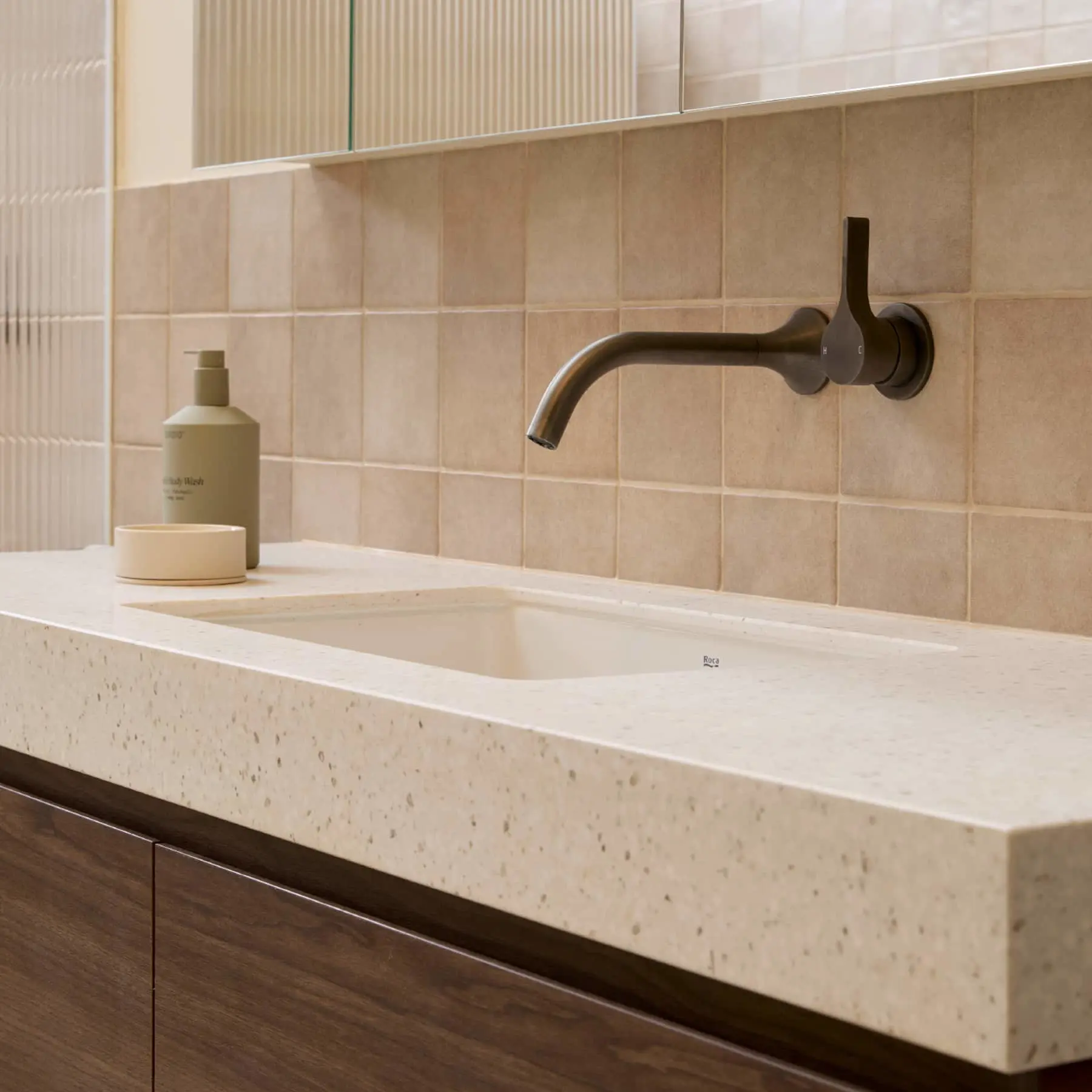 Modern bathroom sink with stone countertop, wall-mounted black faucet, and beige soap dispenser against tiled wall.