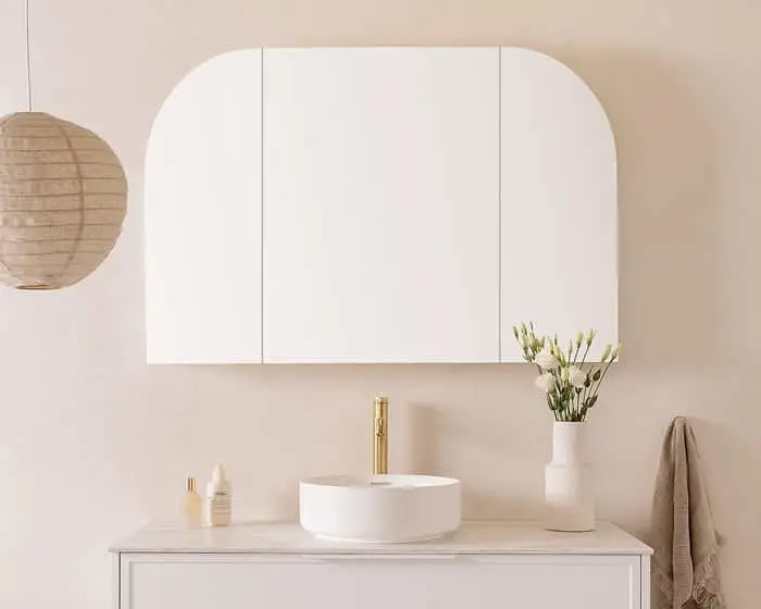 Modern bathroom with white cabinet, round sink, gold faucet, paper lantern, and white flowers in vase.