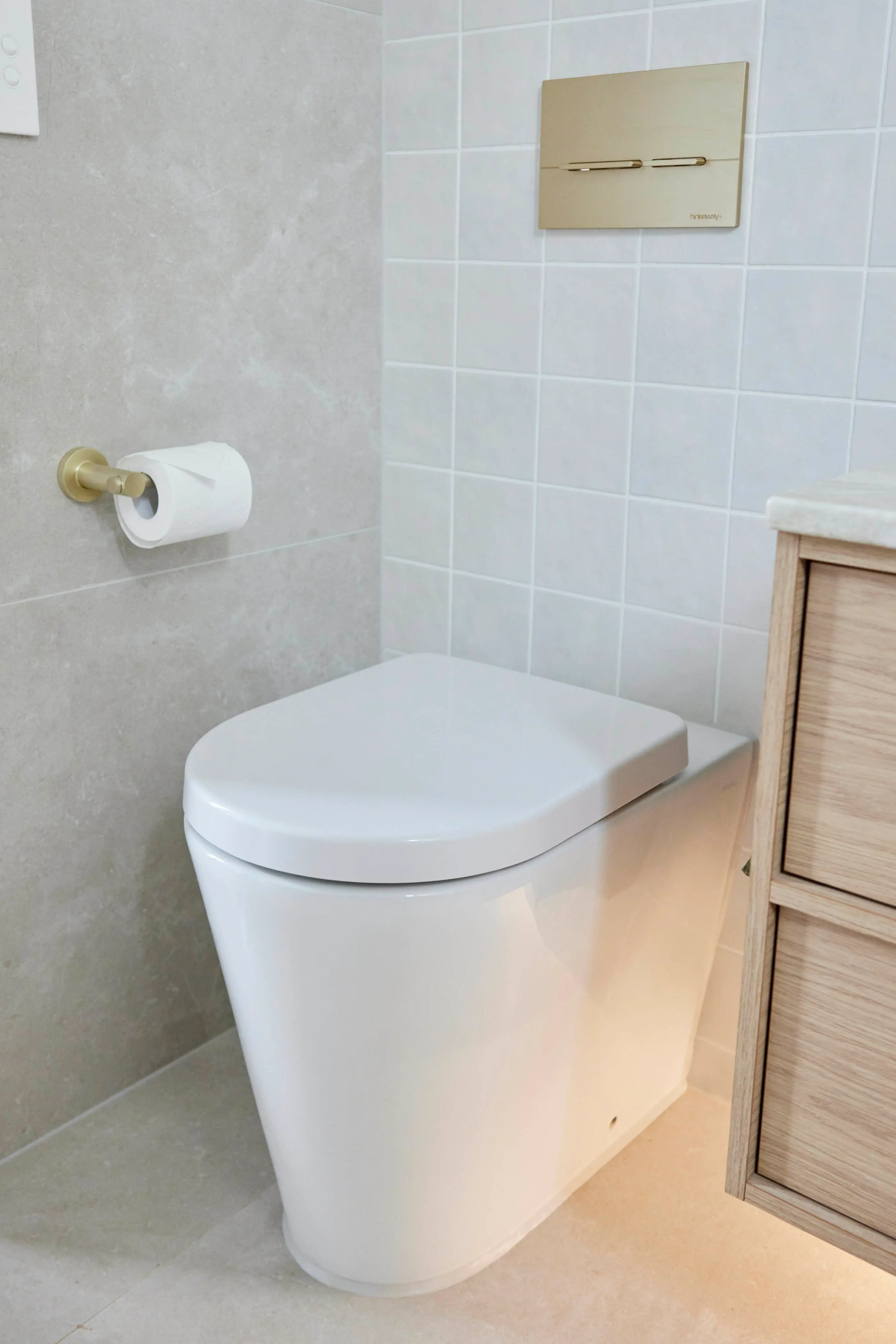 Modern white toilet with gold flush plate, toilet paper holder, and light blue tiled wall in a minimalist bathroom.