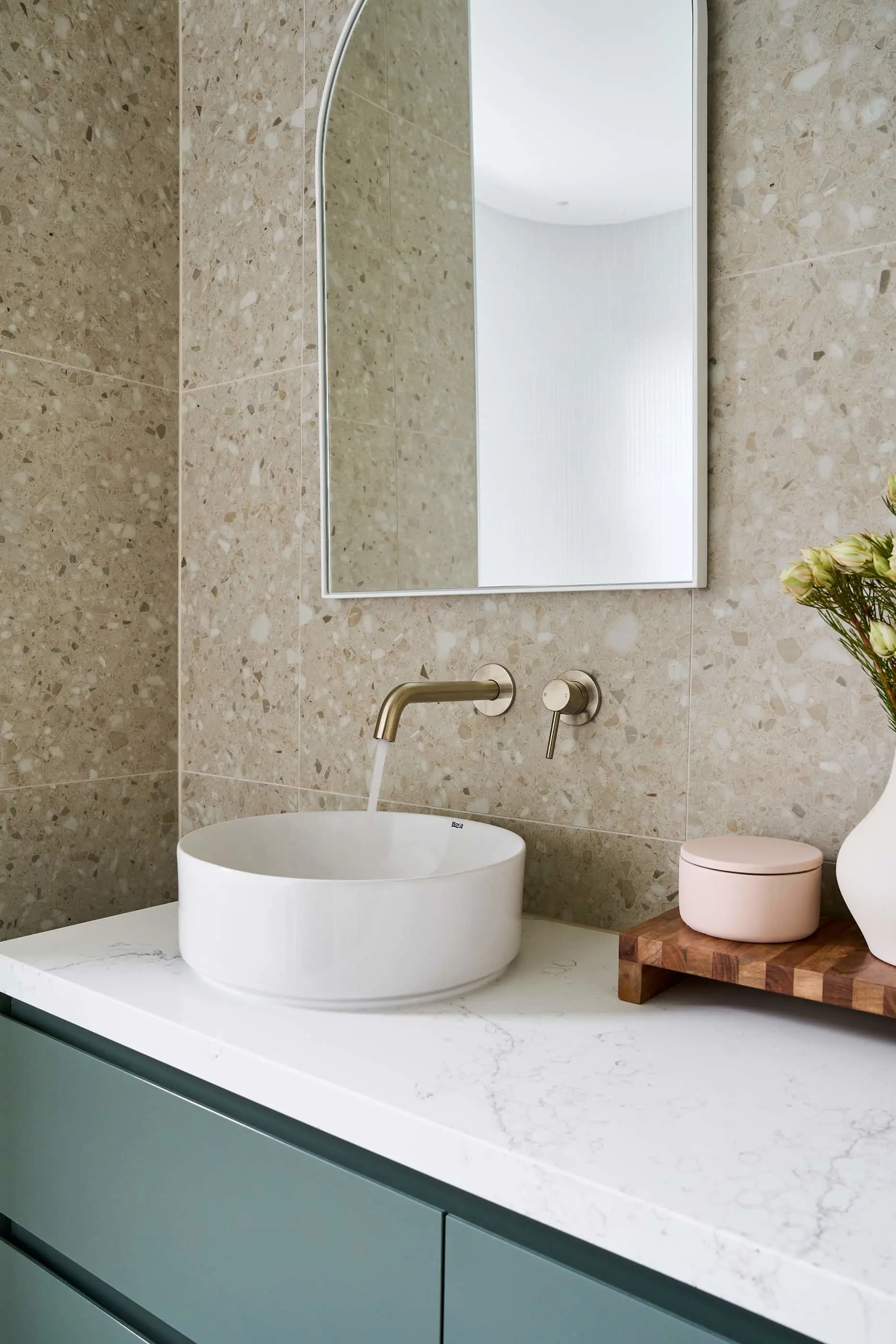 Modern bathroom with terrazzo walls, white vessel sink, brass wall-mounted faucet, and rectangular mirror above teal vanity.