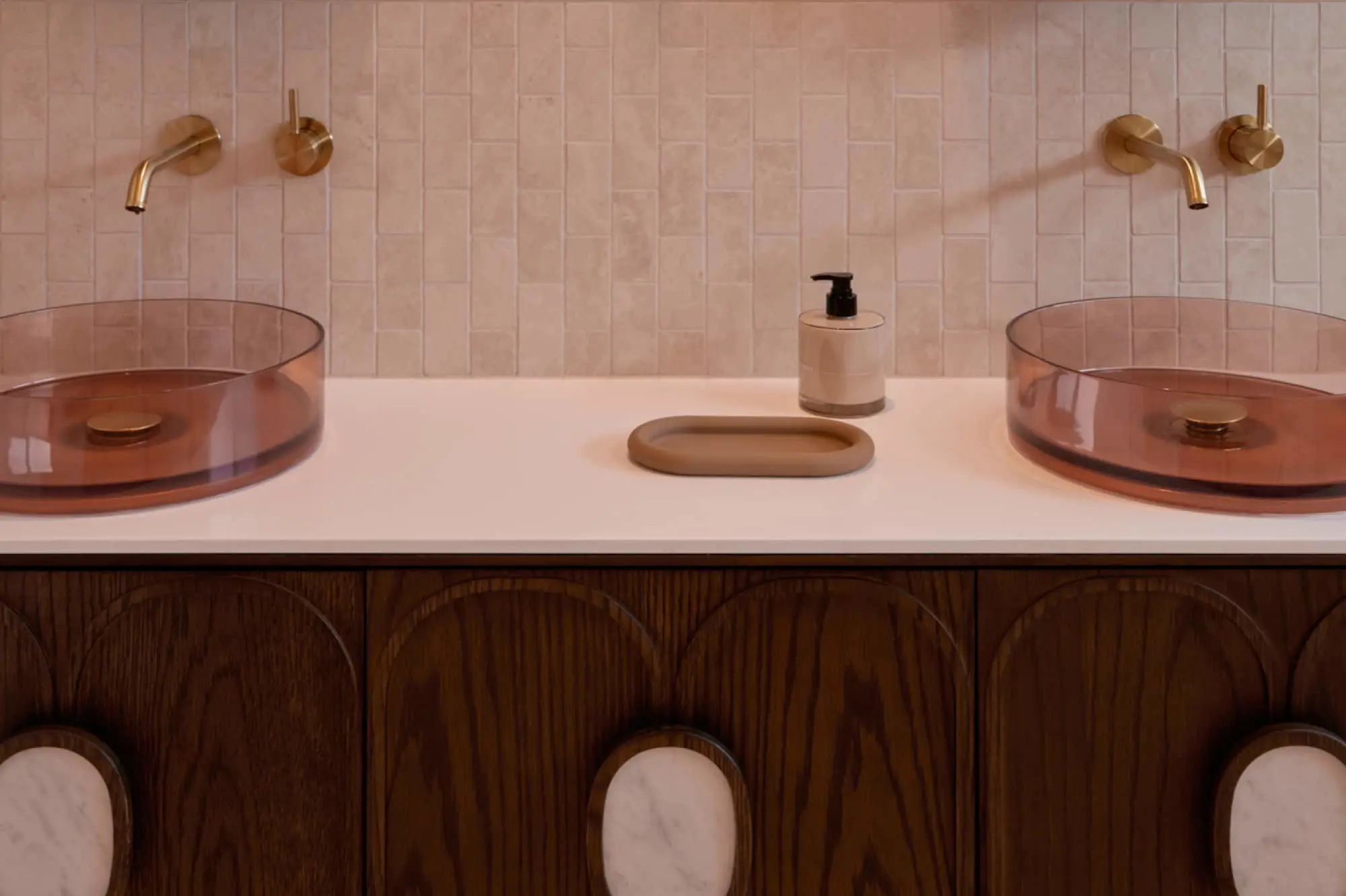 Modern bathroom vanity with dual amber glass sinks, brass wall-mounted faucets, and cream tile backsplash on wooden cabinetry.
