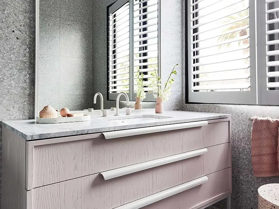 Modern bathroom vanity with marble countertop, white shuttered windows, and pink accents including small vase and towel.