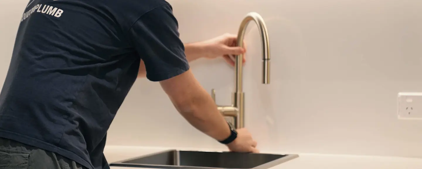 Plumber in navy shirt installing a modern stainless steel kitchen faucet over a sink against white countertop.
