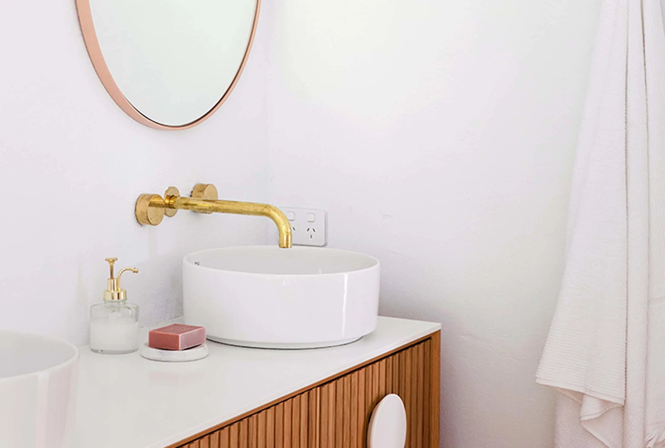 Modern bathroom with white vessel sink, brass wall-mounted faucet, wooden vanity, and round copper-framed mirror.