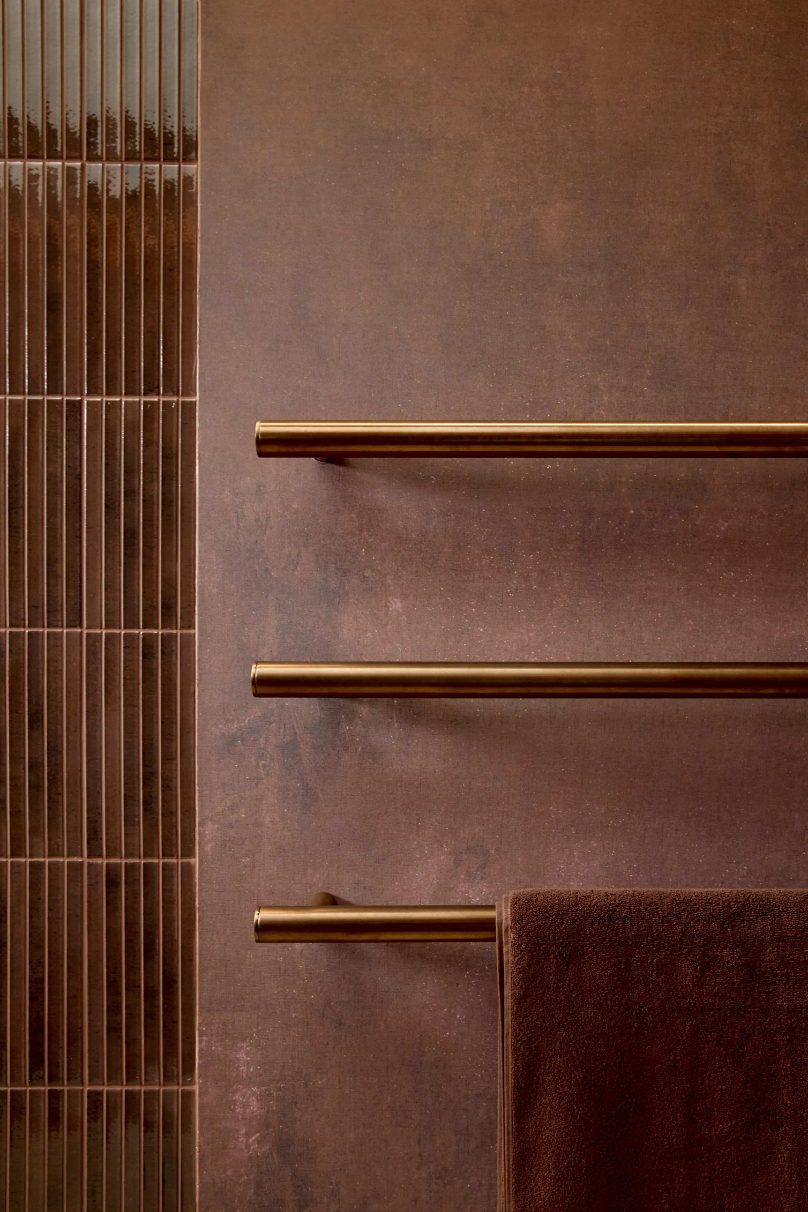 Three brass towel bars mounted on a brown textured wall with vertical tile panels and a brown towel hanging.