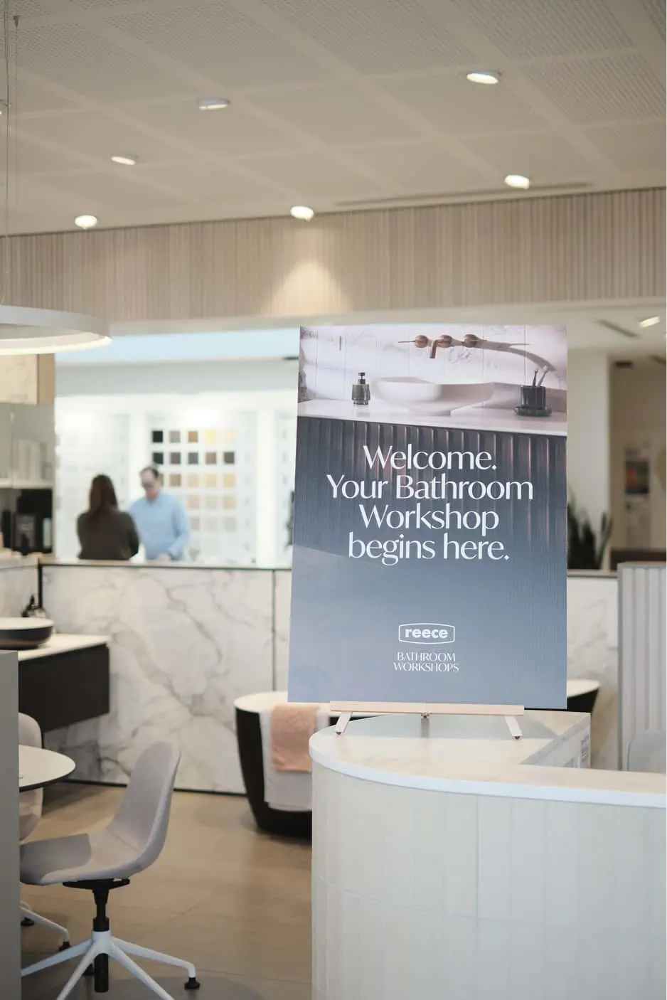 Modern bathroom showroom with welcome sign for Reece Bathroom Workshops, featuring marble counters and display areas.