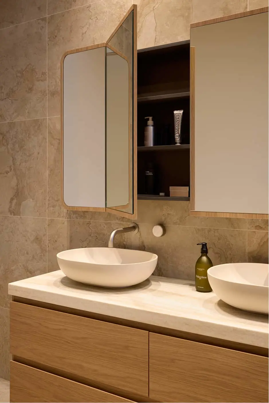 Bathroom vanity with two white vessel sinks, wood cabinet, open mirror cabinet with shelves, and beige tile walls.