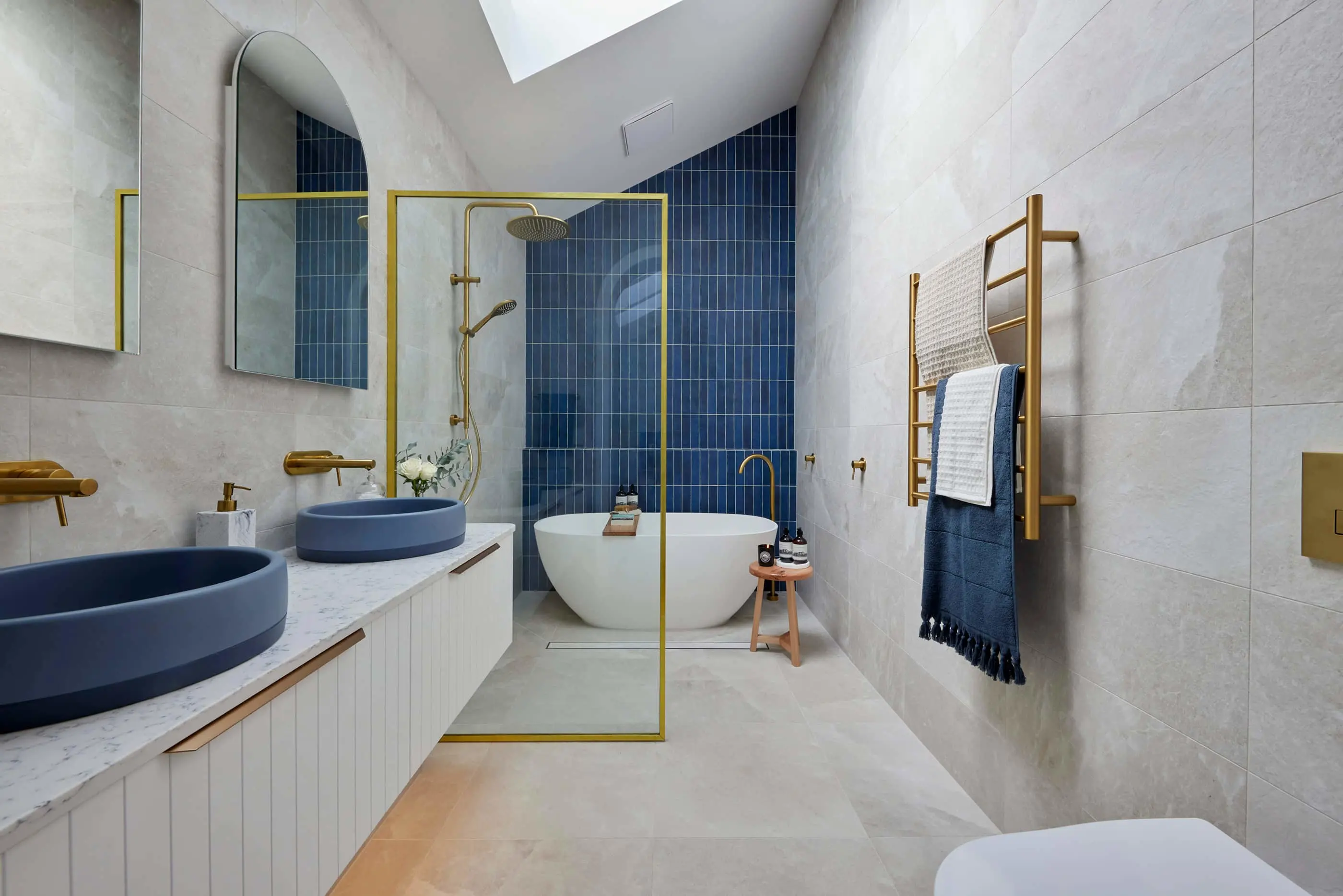 Modern bathroom with blue accent wall, white freestanding tub, gold fixtures, blue sinks, and brass towel rack.