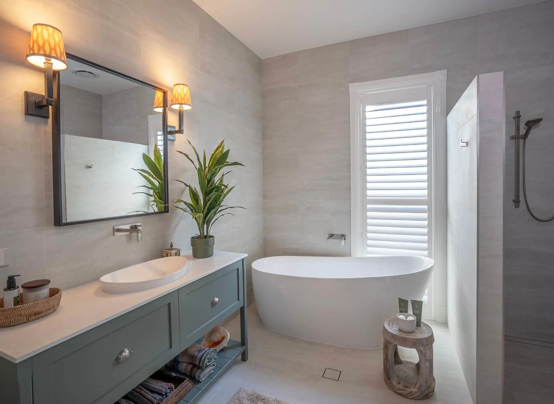 Modern bathroom with blue-green vanity, white freestanding tub, potted plant, and shuttered window.