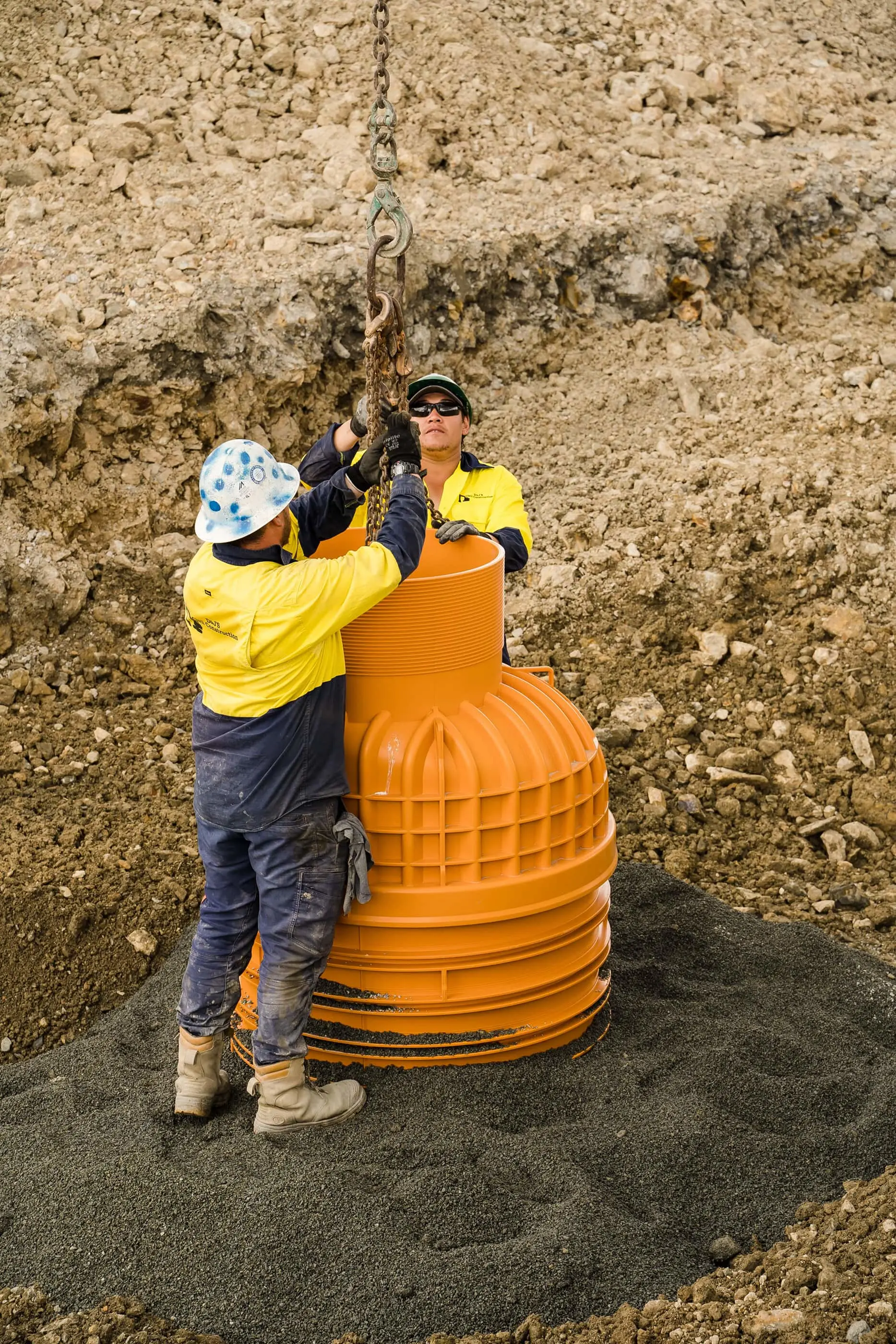 Two workers in yellow safety shirts installing an orange drainage pipe in an excavated dirt area using a chain hoist.