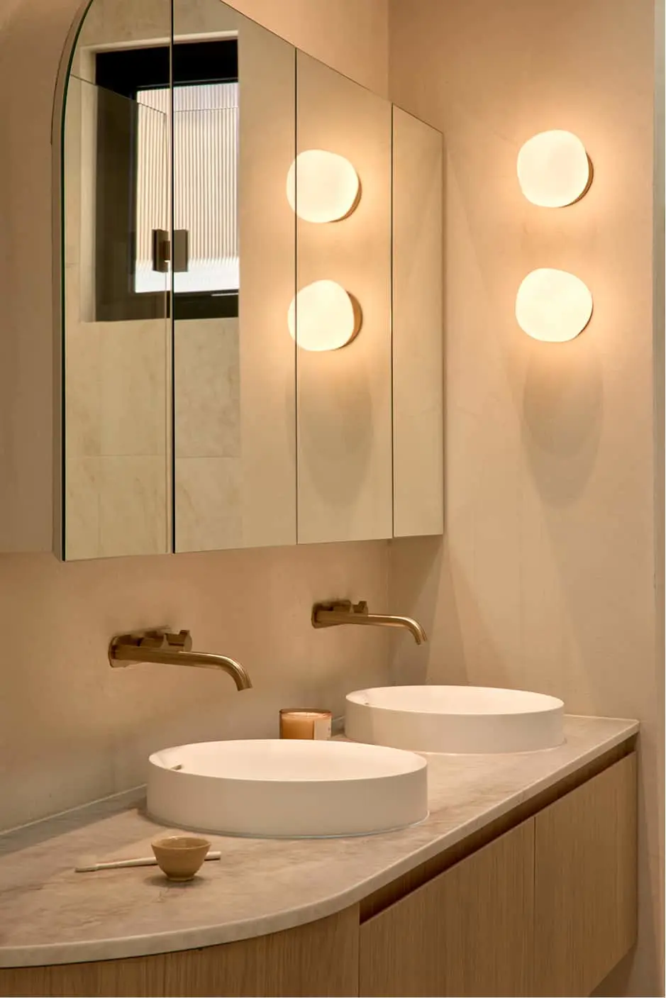Modern bathroom with double white vessel sinks, brass wall-mounted faucets, mirror cabinet, and two round wall lights.