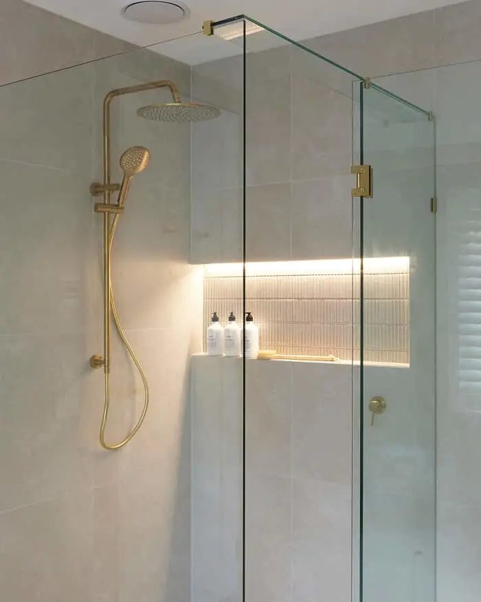 Modern glass shower with gold fixtures, beige tiled walls, and illuminated recessed shelf holding toiletry bottles.