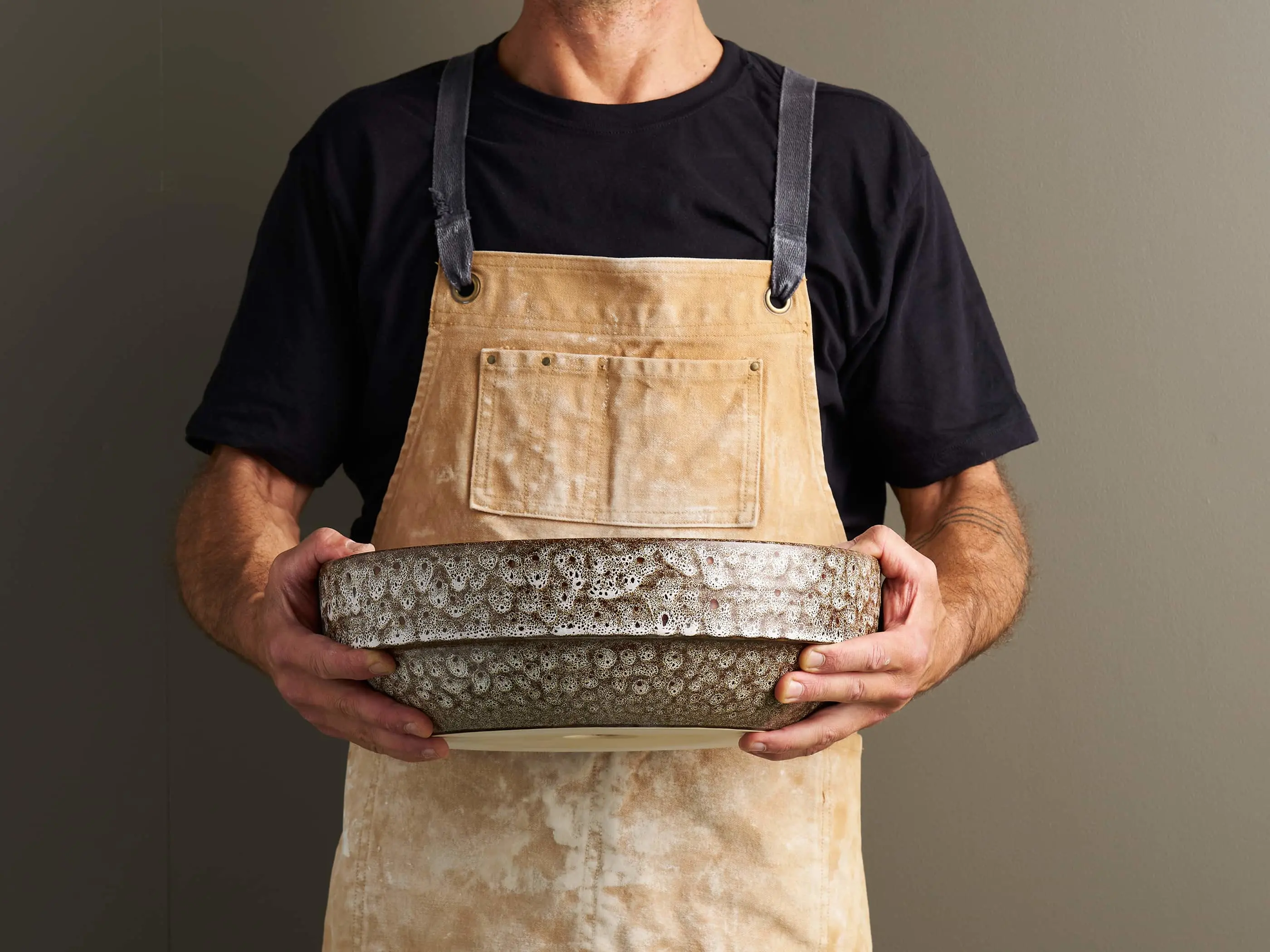A man in a black t-shirt and tan canvas apron holds a large textured ceramic bowl with a speckled glaze.