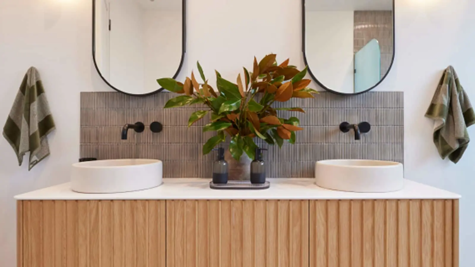 Modern bathroom vanity with dual white vessel sinks, wooden cabinet, gray tile backsplash, round mirrors, and potted plant.