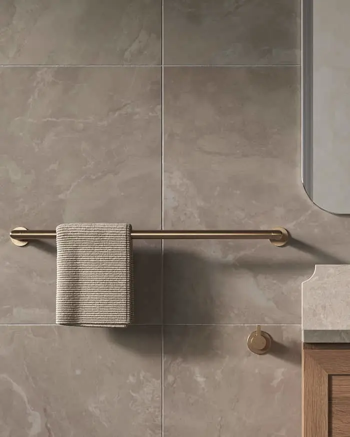 A brass towel bar mounted on gray stone tiles holds a striped gray towel, with a mirror and vanity visible nearby.