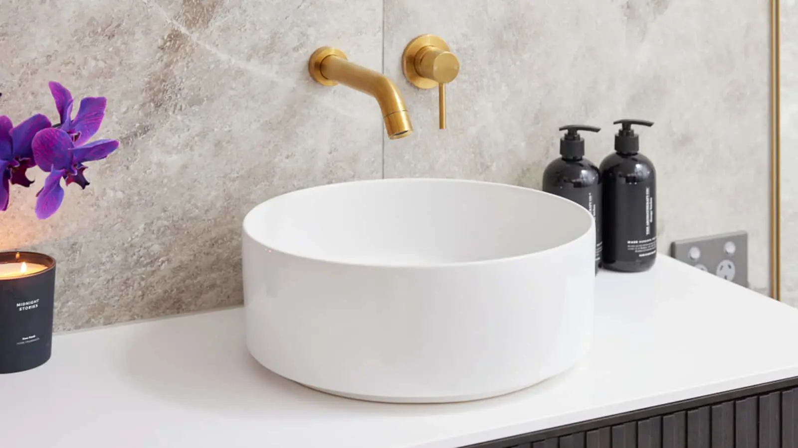 Modern bathroom with white vessel sink, gold wall-mounted faucet, purple orchids, and black soap dispensers.