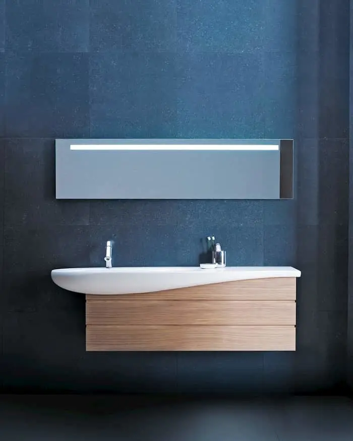 Modern bathroom vanity with wooden cabinet, white sink, and illuminated rectangular mirror against dark blue tiled wall.