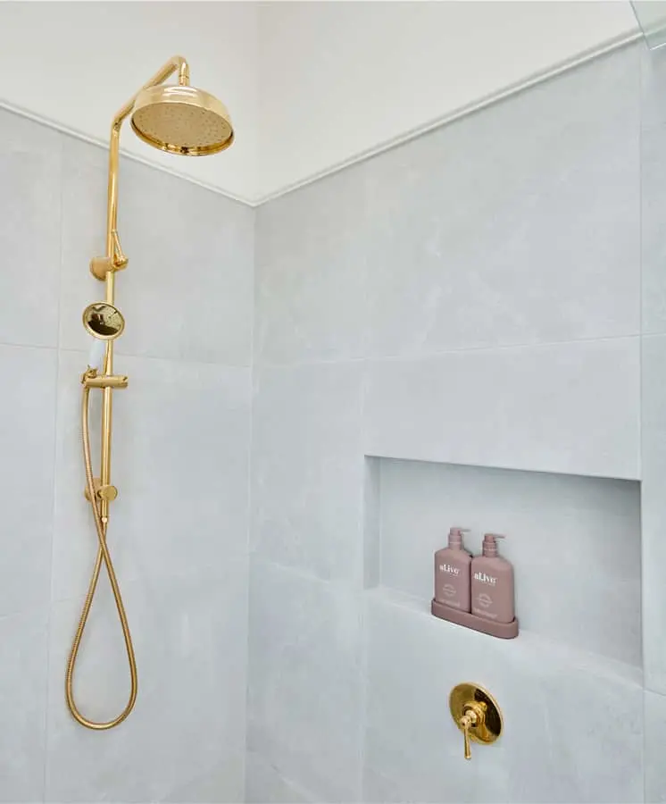 Modern bathroom shower with gold fixtures, rainfall showerhead, handheld sprayer, and built-in niche holding pink bottles.
